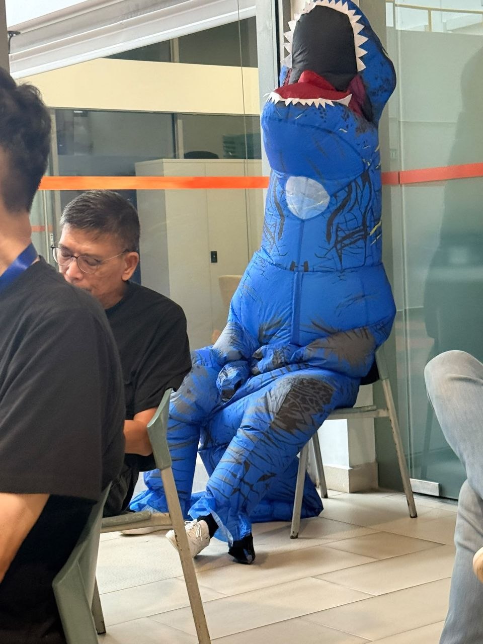 A person in an inflatable T-rex costume sitting in an office — a humorous take on someone who got restructured out of a job